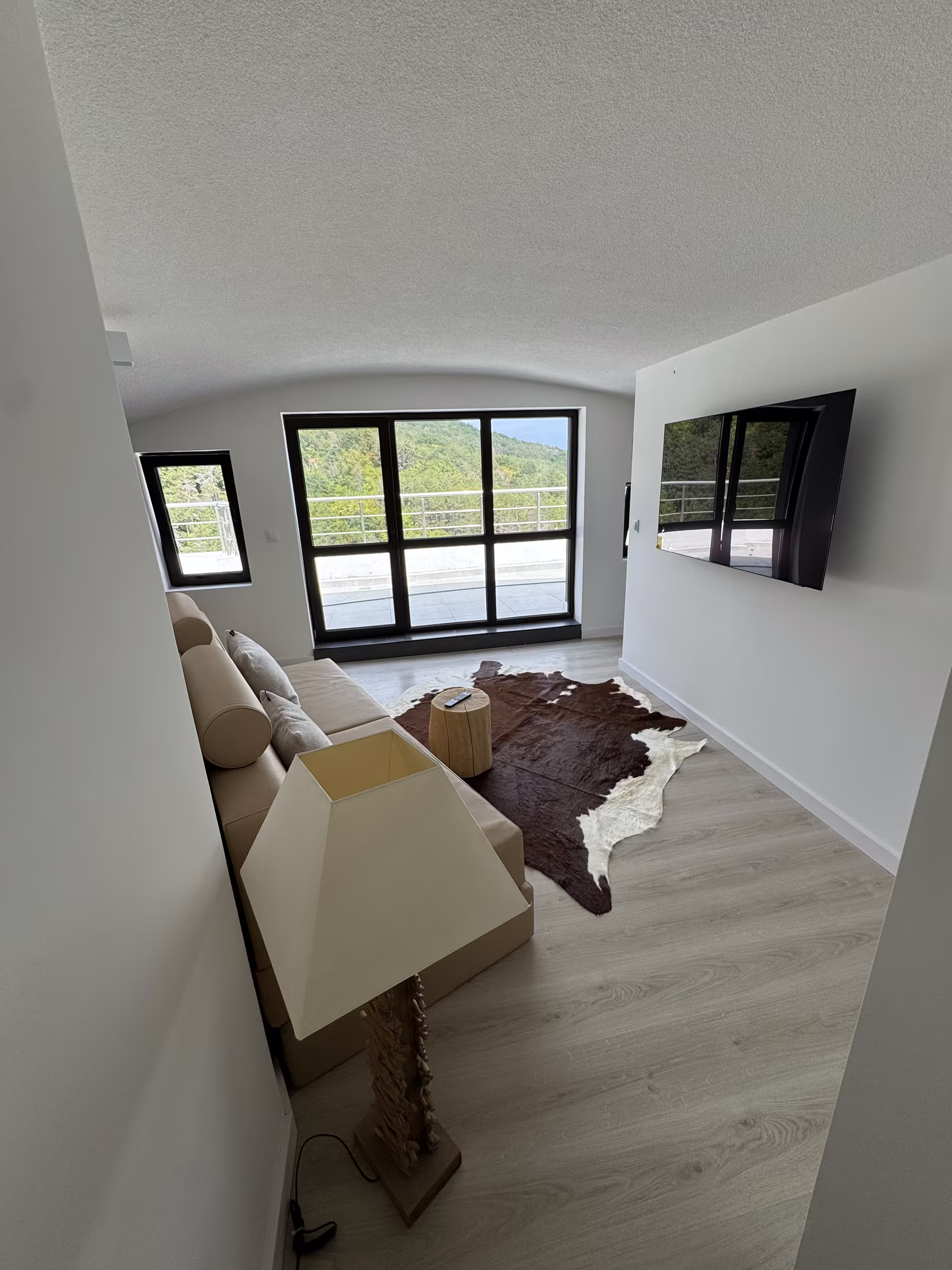 Wohnraum mit Sofa vor der Fensterfront, dahinter Gr&uuml;n und Himmel