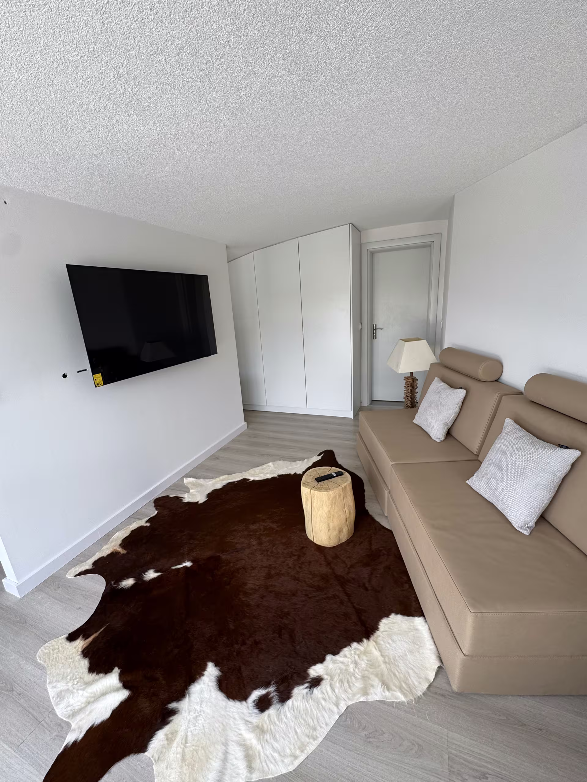 Wohnraum mit Fernseher an der Wand, Ecksofa und Kuhfell-Teppich auf hellem Holzboden