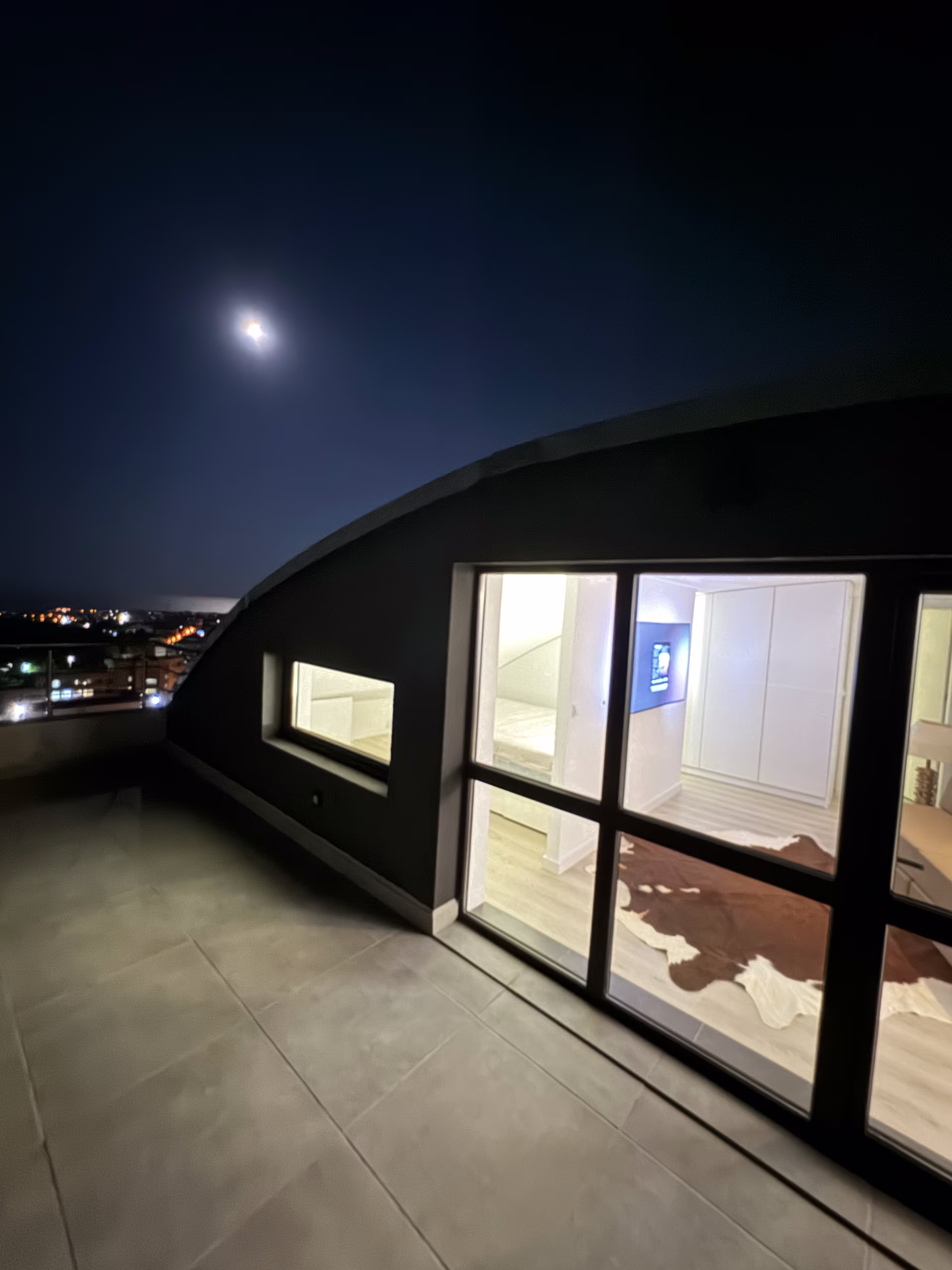 Gefliste Dachterrasse am Abend, der Mond &uuml;ber der geschwungenen Fensterfront, unten die Lichter von Varna