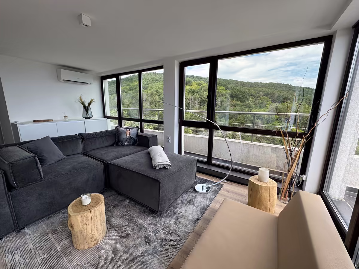 Sofa und Couchtisch, dahinter bewaldete H&uuml;gel